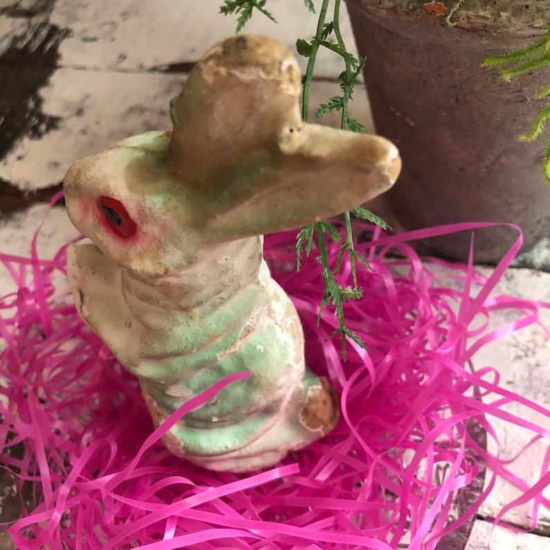 May include: A vintage-style rabbit figurine featuring a weathered, pale green and white finish with a textured surface. The rabbit has prominent ears, a rounded body, and a single visible red eye, displaying signs of age and wear on its exterior.