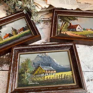 May include: Three framed miniature paintings of countryside scenes. Each painting features a small house, trees, and a blue sky. The paintings are in brown frames with a white distressed finish. The artist's signature, "Canice", is visible in the bottom right corner of each painting.