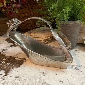 May include: A vintage silver-toned basket with a handle, featuring a grape cluster design on one end. The basket has an elongated shape and sits on a distressed white wooden surface. The background includes flowers and greenery.