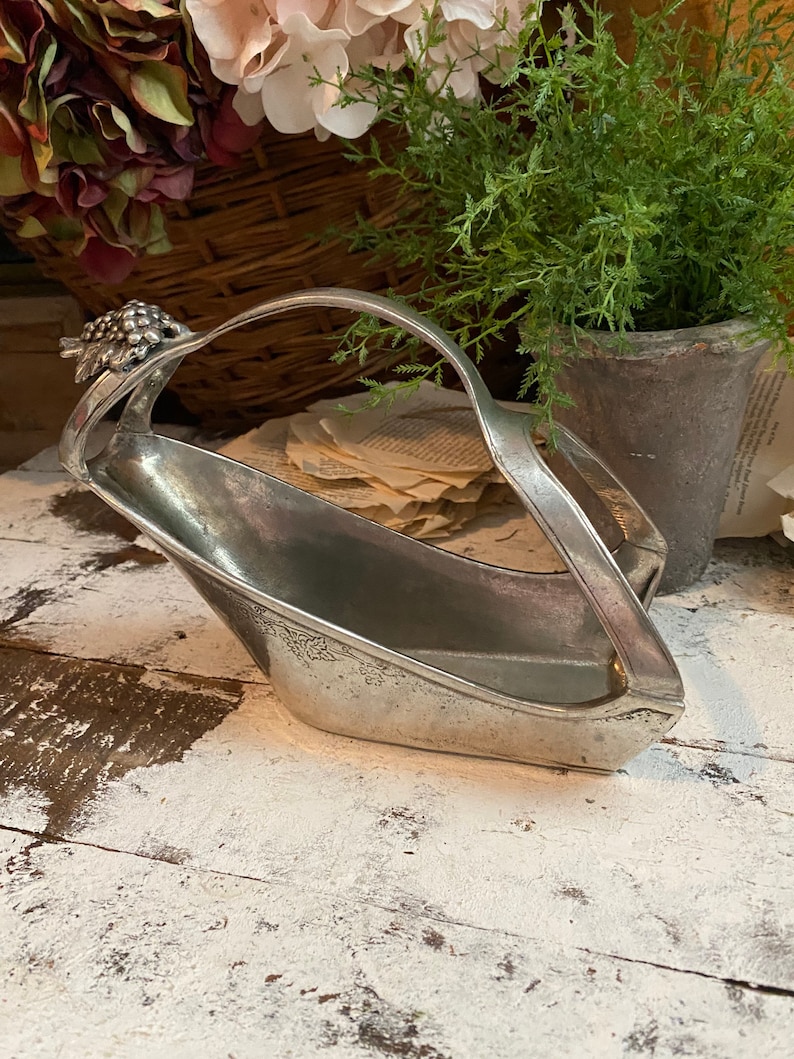 May include: A vintage silver-toned basket with a handle, featuring a grape cluster design on one end. The basket has an elongated shape and sits on a distressed white wooden surface. The background includes flowers and greenery.