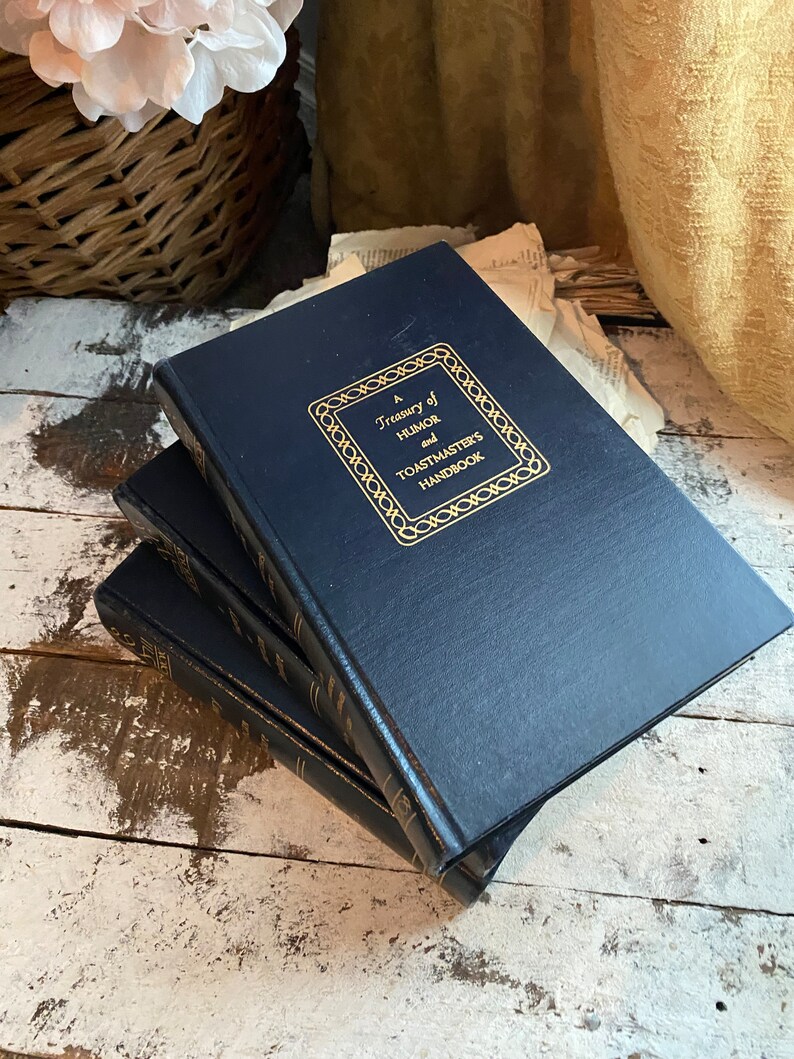 Puede incluir: Una pila de tres libros de tapa dura de color azul marino con letras doradas. El libro de arriba tiene el t&iacute;tulo "A Treasury of Humor and Toastmasters Handbook".