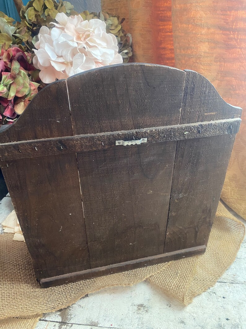 Pu&ograve; includere: Un ripiano a muro in legno marrone scuro con la parte superiore curva e un gancio di metallo sul retro. Il ripiano &egrave; appoggiato su una superficie di juta.