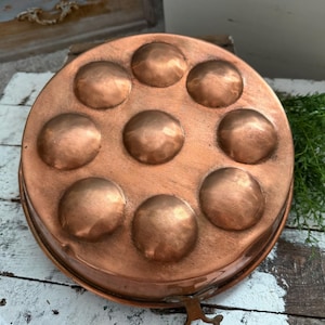 May include: A copper escargot pan with nine circular indentations. The pan is round with a handle and a rich, warm copper color. The pan is designed for cooking and serving escargots.