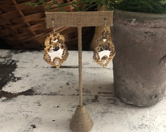 May include: A pair of gold filigree earrings with a mirror-like finish. The earrings are displayed on a brown burlap earring stand.