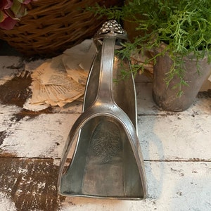 May include: A silver-toned wine bottle holder with a handle and a grape cluster design at the top. The holder has a curved shape with a leaf pattern inside. The background includes plants and a rustic wooden surface.