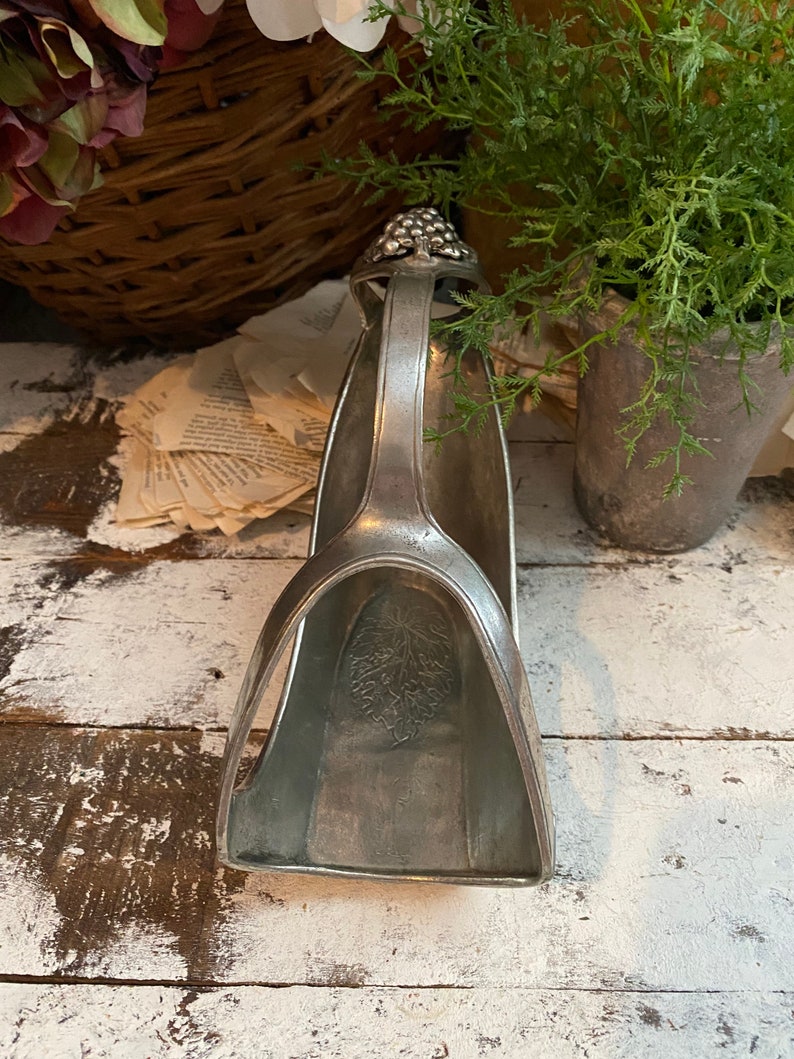 May include: A silver-toned wine bottle holder with a handle and a grape cluster design at the top. The holder has a curved shape with a leaf pattern inside. The background includes plants and a rustic wooden surface.