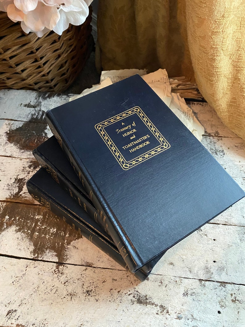 Puede incluir: Tres libros de tapa dura de color azul marino apilados sobre una superficie de madera blanca. El libro de arriba tiene letras doradas que dicen "A Treasury of Humor and Toastmasters Handbook".