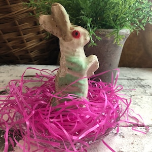 May include: A vintage-style rabbit figurine with a distressed white finish, light green horizontal stripes on its body, and prominent red eyes. The rabbit is standing upright on its hind legs and is nestled within a clear glass bowl filled with bright pink shredded paper filler.