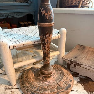 May include: A distressed, antique-style candlestick holder. The holder features a dark brown, peeling paint finish with hints of pink and a decorative base. The candlestick is likely made of wood and has a rustic, vintage aesthetic.