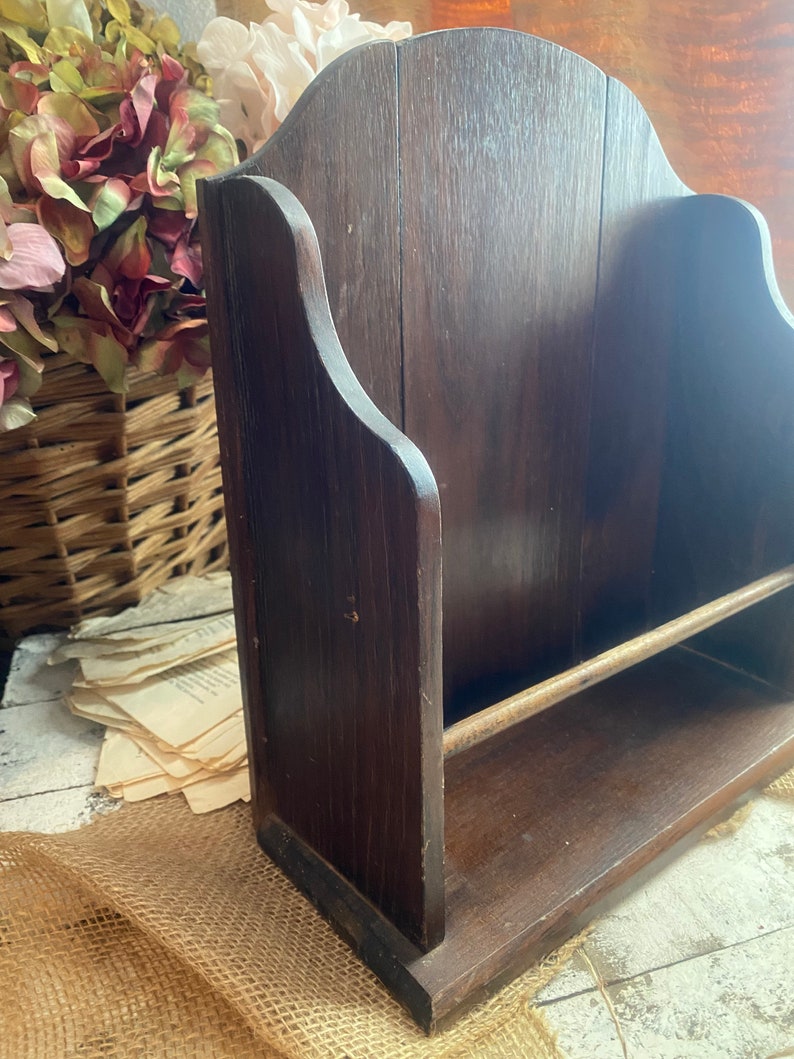 Pu&ograve; includere: Un porta riviste in legno marrone scuro con uno scaffale e un'asta di metallo. Il porta riviste ha una parte superiore curva ed &egrave; fatto di legno con venature naturali.
