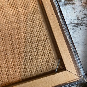 May include: Close-up of the back of a framed picture. The frame is made of wood with a dark brown finish. The back of the picture is made of a woven material. A metal hook is attached to the frame.