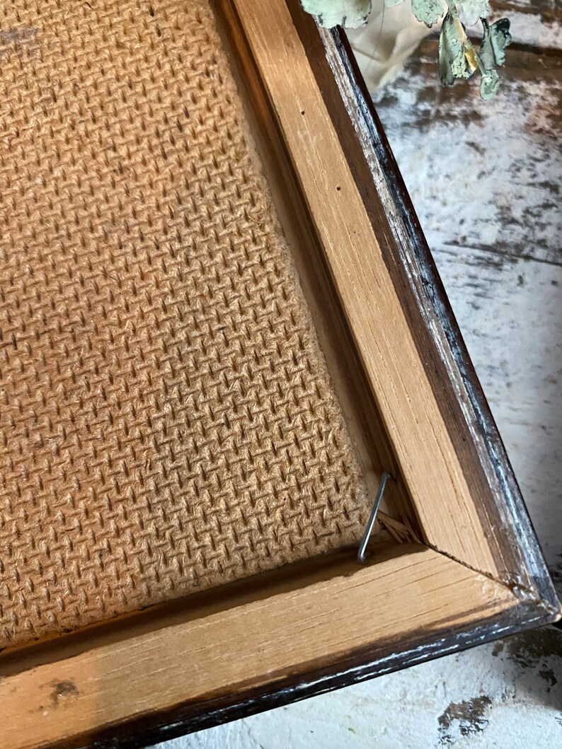 May include: Close-up of the back of a framed picture. The frame is made of wood with a dark brown finish. The back of the picture is made of a woven material. A metal hook is attached to the frame.