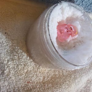 May include: A clear, cylindrical plastic jar filled with a white, granular substance. Embedded within the center of the white material is a small, translucent pink figurine shaped like a pig with subtle facial features and a yellow accent.