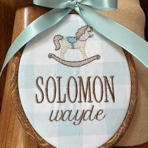 Pode incluir: Um bastidor de bordado oval contendo tecido xadrez azul claro e branco. O tecido apresenta um cavalo de balanço bordado em bege e azul, com o texto SOLOMON wayde em fio castanho. Uma fita de cetim azul claro está atada no topo.