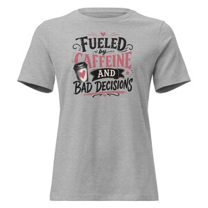 Puede incluir: Una camiseta gris jaspeado de manga corta con un estampado gráfico que incluye el texto "FUELED BY CAFFEINE AND BAD DECISIONS" en letras negras y rosas, acompañado de la ilustración de una taza de café con un pequeño corazón rosa.