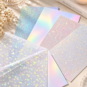 May include: A collection of rectangular sheets featuring various holographic finishes, including a blue glitter pattern, a smooth iridescent gradient, a pink star-speckled texture, and a clear film with reflective geometric shards.