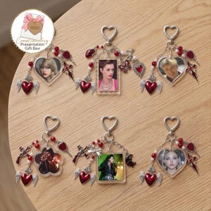 Puede incluir: Seis llaveros decorativos con marcos de acrílico en forma de corazón o rectangulares que contienen retratos de celebridades. Cada llavero incluye un cierre de corazón, cuentas rojas y blancas, un corazón alado rojo y dijes metálicos adicionales como cruces o mariposas.