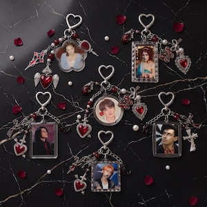 May include: Five decorative keychains featuring heart-shaped clasps, each displaying a different portrait within a frame. The charms include red enamel hearts, silver-toned crosses, butterflies, and small white beads, creating a cohesive aesthetic.