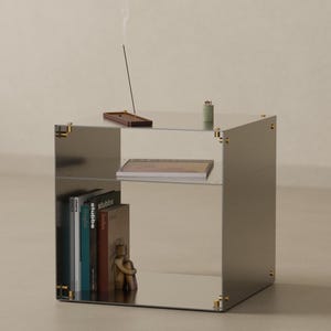 May include: A square, mirrored side table with a reflective surface and gold-toned corner connectors. The table features two open shelves, with the bottom shelf holding several books and a small figurine, and the top shelf holding a book.