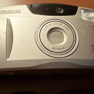 May include: A silver Canon Sure Shot 76 Zoom point-and-shoot film camera with a 38-76mm lens. The front features the Canon logo, a built-in flash, and a black viewfinder window. A black wrist strap is attached to the side of the camera body.