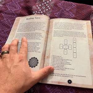 Może przedstawiać: Otwarta książka z brązową okładką i brązowymi stronami. Książka nosi tytuł "Reading Tarot" i zawiera schemat rozkładu kart tarota z ponumerowanymi pozycjami i pytaniami dla każdej pozycji. Książka jest otwarta na stronie 4.