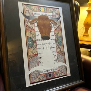 May include: A framed print featuring a brown bull's head illustration in the centre. The print has a decorative border with intricate patterns and the text "when the two bills caught... in a blacksmith's forge."