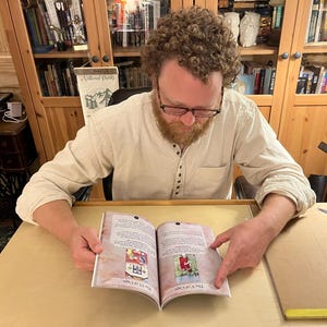 Może przedstawiać: Osoba czyta książkę z kolorowymi ilustracjami. Książka jest otwarta na stronie z tekstem "The VII of Cups" i "The V of Cups".