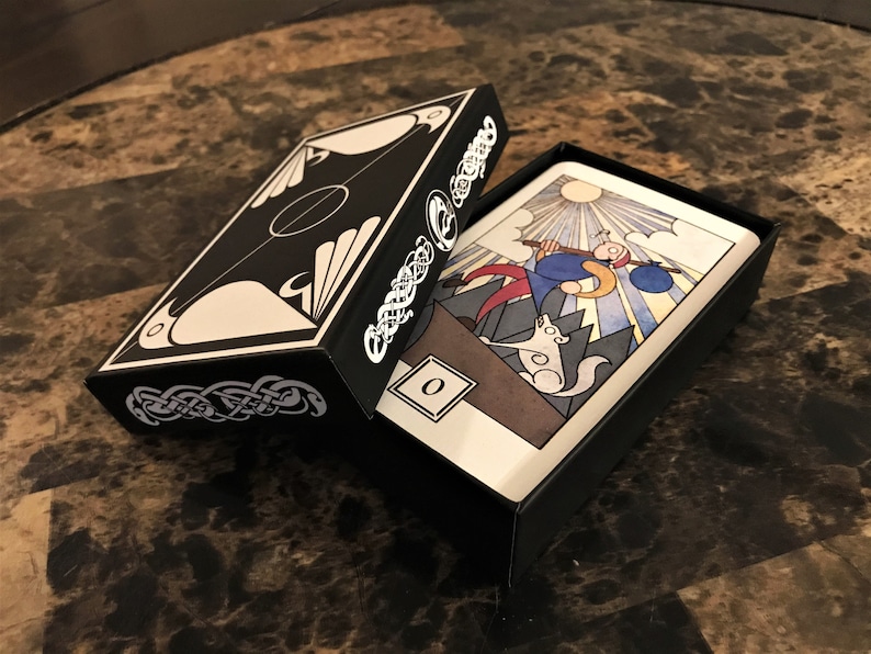 May include: A black box with a white Celtic knot design and a white and black illustration of a bird on the lid. The box is open and a tarot card with a colorful illustration of a person walking with a dog is visible. The card has the number "0" in the bottom left corner.