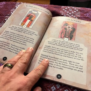 Może przedstawiać: Otwarta książka z dwoma stronami przedstawiającymi ilustracje kart tarota. Karta Hierofanta przedstawia postać w czerwonej szacie i czarnym kapeluszu stojącą w drzwiach. Karta Zakochanych przedstawia dwie postacie z czarnymi skrzydłami i wężem splecionym z ich ciałami. Tekst na stronach brzmi "Hierofanta" i "Zakochanych".