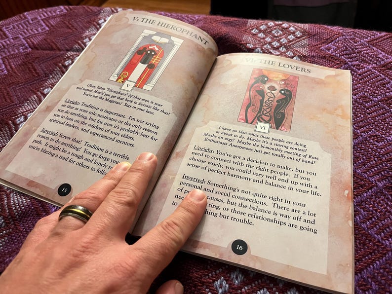 Może przedstawiać: Otwarta książka z dwoma stronami przedstawiającymi ilustracje kart tarota. Karta Hierofanta przedstawia postać w czerwonej szacie i czarnym kapeluszu stojącą w drzwiach. Karta Zakochanych przedstawia dwie postacie z czarnymi skrzydłami i wężem splecionym z ich ciałami. Tekst na stronach brzmi "Hierofanta" i "Zakochanych".