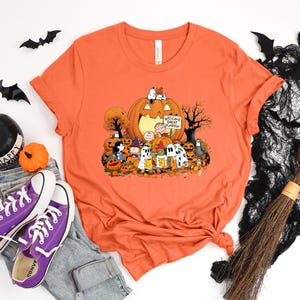 May include: An orange short-sleeved t-shirt featuring a graphic print of the Peanuts characters celebrating Halloween. The design includes Snoopy, Charlie Brown, and friends surrounded by pumpkins and a sign that reads Welcome Great Pumpkin.