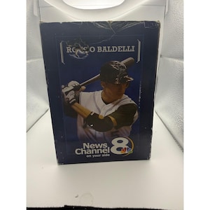 May include: A rectangular cardboard box featuring a graphic of baseball player Rocco Baldelli in a white and black uniform holding a bat. The dark blue packaging displays the text Rocco Baldelli and the News Channel 8 on your side logo.