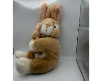 May include: A plush stuffed animal featuring a large tan rabbit with white paws and inner ears, holding a smaller matching tan baby rabbit in its arms. The rabbits have soft fur, dark eyes, and stitched noses, presented in a seated position.