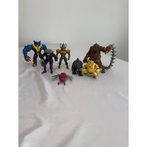 May include: A collection of six plastic action figures including a blue and yellow hero, a figure in a black coat, a gold armored warrior, a small black creature, a yellow sumo-style character, and a brown monster with a spiked tail.