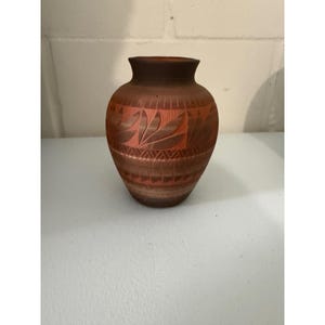 May include: A terracotta-colored vase with a rounded body and a narrow neck. The surface features carved geometric patterns, including a band of stylized leaf shapes near the top and a series of horizontal lines and chevron motifs around the center.