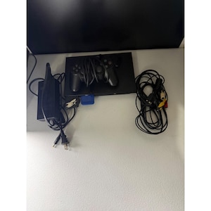 May include: A black video game console with a matching controller resting on top. The set includes a black power adapter with a cord and plug, and a bundle of black AV cables with yellow, white, and red connectors. A small blue memory card is inserted.