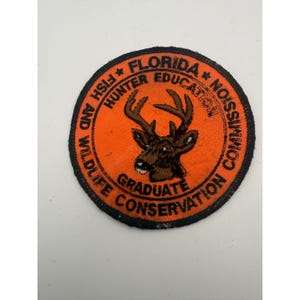 May include: A circular embroidered patch with a bright orange background and a black border. In the center is a brown deer head with antlers. The text reads FLORIDA FISH AND WILDLIFE CONSERVATION COMMISSION, HUNTER EDUCATION, and GRADUATE.