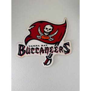May include: A Tampa Bay Buccaneers logo decal featuring a red flag with a white skull and crossbones, a brown football, and the text Tampa Bay Buccaneers in black and red lettering with a sword graphic extending from the bottom of the flag.