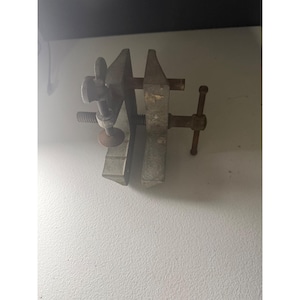 May include: A metal bench vise featuring two parallel jaws with a textured gray finish. The tool includes a threaded screw mechanism with a T-shaped handle for adjustment and a secondary vertical wing nut clamp attached to the side of the main body.
