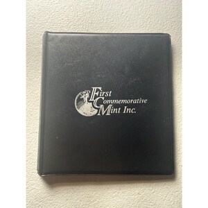 May include: A square black binder with a textured finish featuring the silver-colored logo and text First Commemorative Mint Inc. printed on the front cover. The logo includes a circular graphic depicting a figure standing on a pedestal.