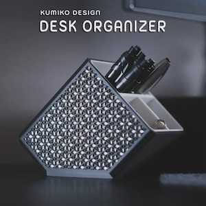May include: A Kumiko Design desk organiser featuring a dark grey exterior with a white interior and a geometric lattice pattern on the front panel. The container has multiple compartments holding black pens and a silver metal binder clip.