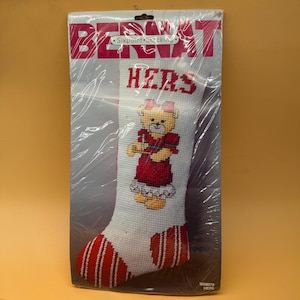 May include: A Bernat Sixpoint Stocking needlepoint kit in original plastic packaging. The white stocking features a cross-stitched teddy bear wearing a red dress and bow, with the word HERS in red letters at the top and red striped accents.