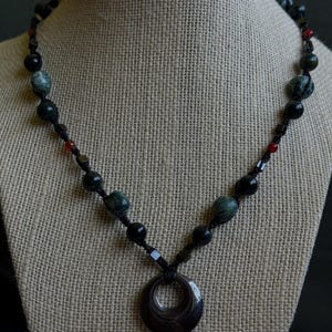 May include: A necklace featuring a dark brown circular pendant suspended from a black cord. The cord is accented with alternating black beads, mottled green stone beads, small metallic cube spacers, and tiny red beads arranged in a symmetrical pattern.