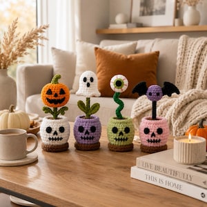 Könnte beinhalten: Vier gehäkelte Halloween-Dekorationen in totenkopfförmigen Töpfen. Die erste zeigt einen orangefarbenen Kürbis, die zweite ein weißes Gespenst, die dritte einen grünen Augapfel auf einem Stiel und die vierte eine lila Fledermaus.