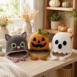 May include: Three crocheted hats featuring Halloween themes: a gray cat with green eyes and pink ears, an orange jack-o'-lantern with a green stem, and a white ghost with black eyes and a mouth. Each hat includes braided earflaps with tassels.