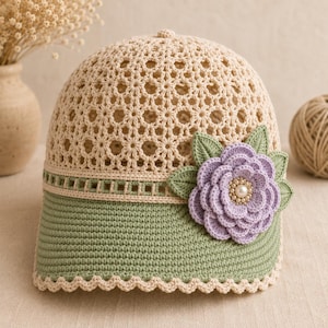 May include: A crochet baseball cap featuring a beige openwork crown, a sage green brim with a scalloped edge, and a decorative lavender flower appliqué with green leaves and a central cluster of small white pearls attached to the side.