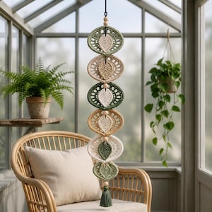 May include: A vertical wall hanging featuring six circular crochet motifs in shades of sage green, cream, and tan. Each motif contains a central leaf-shaped element, separated by small wooden beads, and finished with a green tassel at the bottom.