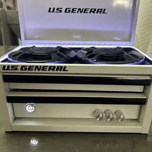Può includere: Una cassetta degli attrezzi bianca U.S. General modificata in un sistema di altoparlanti con due altoparlanti circolari neri sulla parte superiore, un coperchio incernierato, un pulsante di accensione luminoso e tre manopole argentate.