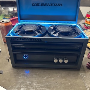 May include: A grey U.S. General metal tool box converted into a speaker system with two black circular speakers on top. The lid is open, revealing blue interior lighting. The front features a blue power button and three silver control knobs.