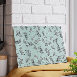 May include: A rectangular glass cutting board featuring a light mint green background with a repeating pattern of black line art illustrations depicting stylized crowns in various orientations and sizes across the entire surface.
